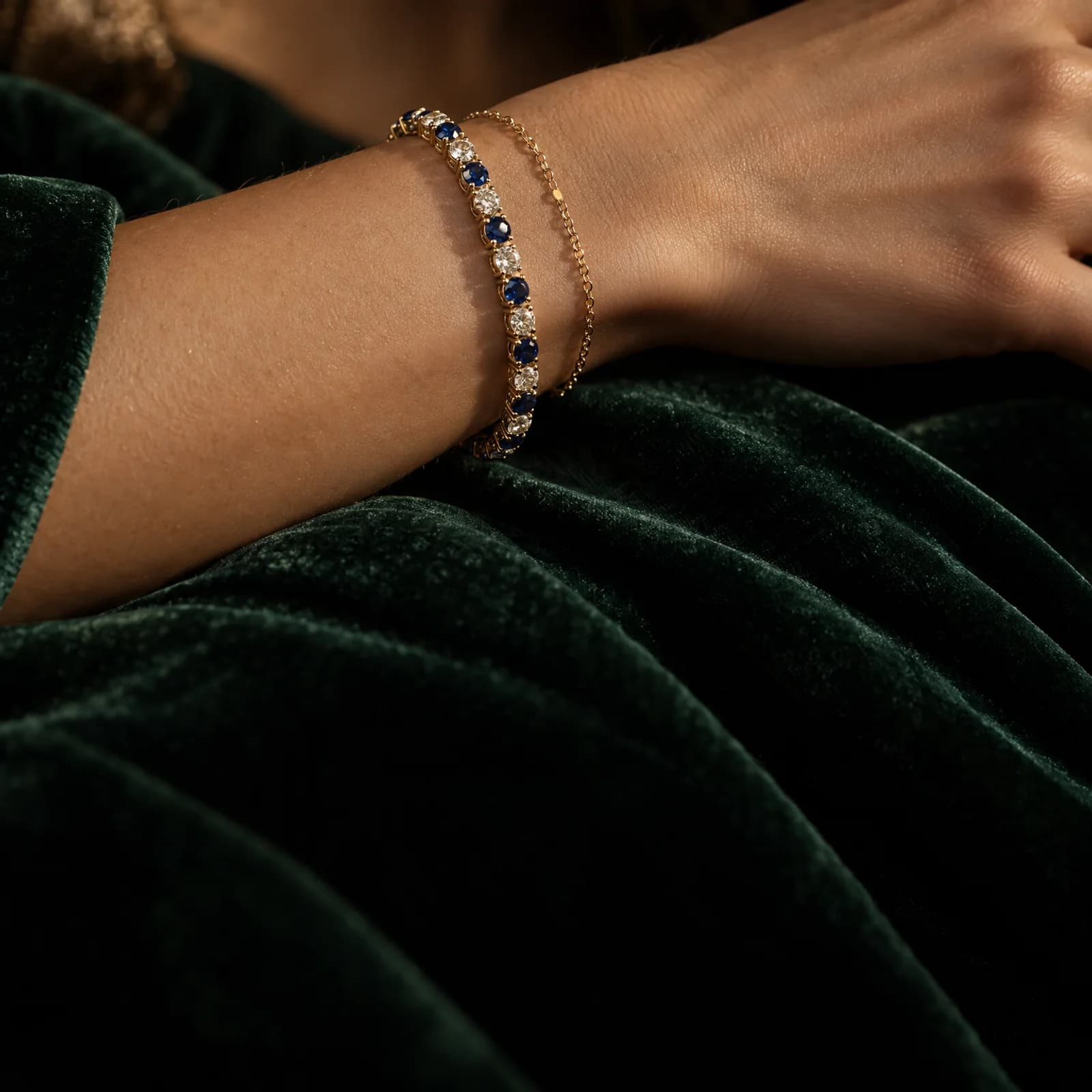 A wrist on green velvet wearing a gold sapphire and diamond tennis bracelet with a fine gold chain